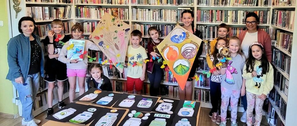 -na zdjęciu widać dzieci stojące w sali bibliotecznej na tle regałów. dzieci trzymają prace plastyczne