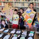 -na zdjęciu widać dzieci stojące w sali bibliotecznej na tle regałów. dzieci trzymają prace plastyczne