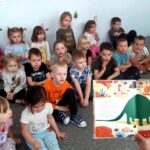 na zdjęciu widać dzieci siedzące na podłodze i słuchające czytanej im książki