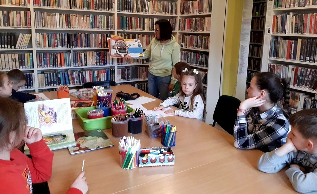 na zdjęciu widać dzieci siedzące wokół stołu w sali bibliotecznej i bibliotekarkę, która pokazuje im książkę