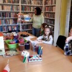 na zdjęciu widać dzieci siedzące wokół stołu w sali bibliotecznej i bibliotekarkę, która pokazuje im książkę