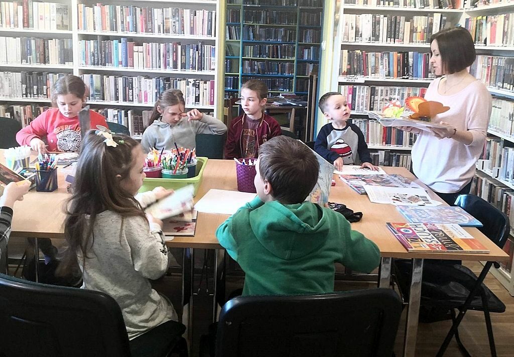 na zdjęciu widać dzieci siedzące wokół stołu w sali bibliotecznej i bibliotekarkę, która pokazuje im książkę