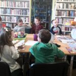 na zdjęciu widać dzieci siedzące wokół stołu w sali bibliotecznej i bibliotekarkę, która pokazuje im książkę