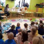 na zdjęciu widać salę przedszkolną oraz dzieci siedzące przed bibliotekarką czytającą im książkę.