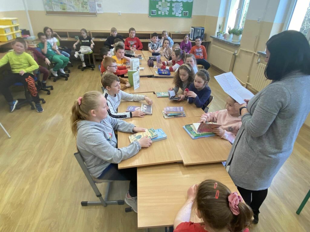 na zdjęciu widać salę szkolną, przy stołach siedzą dzieci, bibliotekarz czyta list skierowany do dzieci
