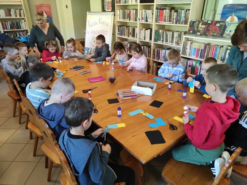 na zdjęciu widać salę biblioteczną, na środku sali widać stół, wokół stołu siedzą dzieci, wykonują pracę plastyczną