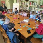 na zdjęciu widać salę biblioteczną, na środku sali widać stół, wokół stołu siedzą dzieci, wykonują pracę plastyczną