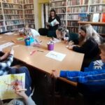 na zdjęciu widać dzieci siedzące przy stole i czytającą im bibliotekarkę.