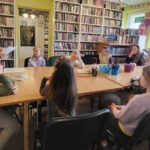 na zdjęciu widać dzieci siedzące przy stole i czytającą im bibliotekarkę.