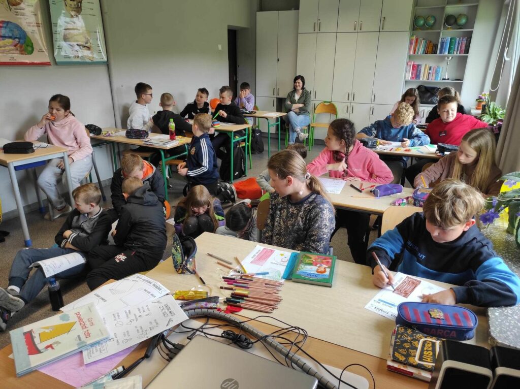 na zdjęciu widać salę szkolną, przy stolikach siedzą dzieci, uzupełnią karty pracy