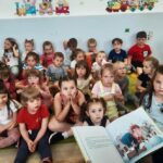 na zdjęciu widać siedzące dzieci, słuchające czytającej im bibliotekarki.