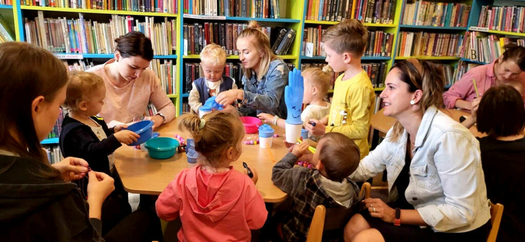 na zdjęciu widać dzieci i ich mamy siedzące przy stolikach w sali bibliotecznej