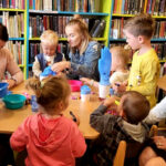 na zdjęciu widać dzieci i ich mamy siedzące przy stolikach w sali bibliotecznej