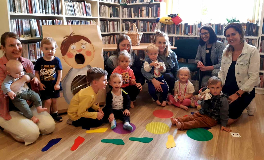 na zdjęciu widać dzieci i mamy siedzące na podłodze w sali bibliotecznej