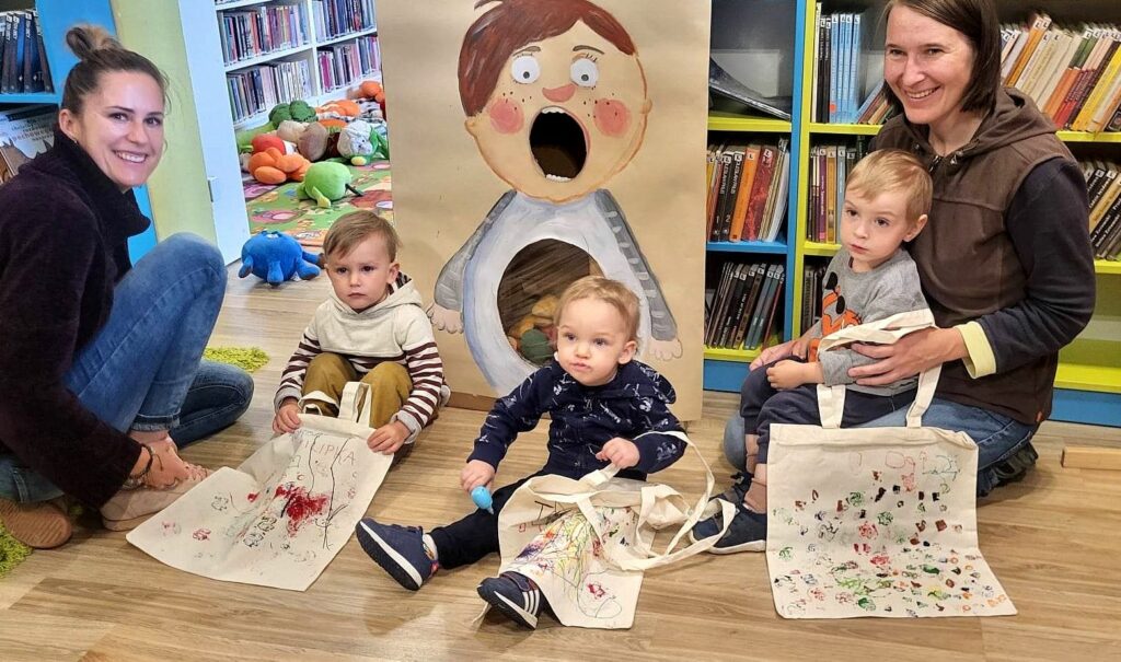 na zdjęciu widać sale biblioteczna oraz małe dzieci wraz z mamami siedzących na podłodze.