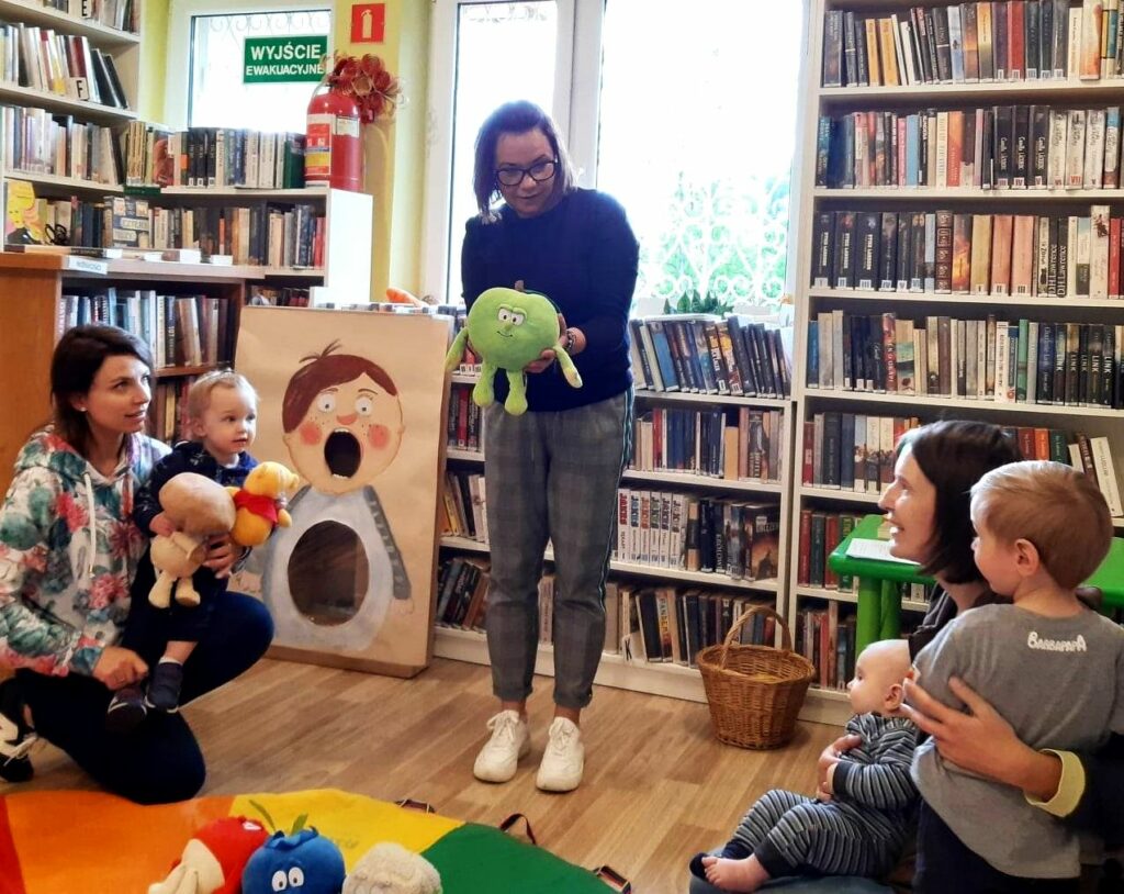 na zdjęciu widać sale biblioteczną oraz małe dzieci z mamami i bibliotekarkę pokazująca im maskotkę.