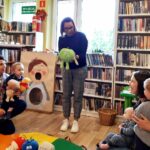 na zdjęciu widać sale biblioteczną oraz małe dzieci z mamami i bibliotekarkę pokazująca im maskotkę.