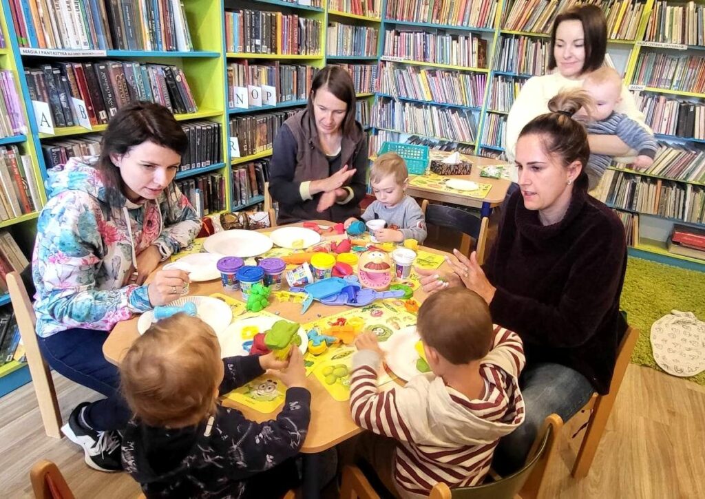 na zdjęciu widać dzieci i mamy siedzące przy stoliku w bibliotecznej sali