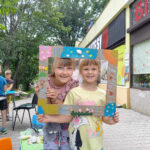 Dwie dziewczynki stoją, przed ich twarzami kolorowa ramka. w tyle przy stole chłopcy wykonują prace plastyczne