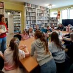 na zdjęciu widać dzieci siedzące wokół stołu w sali bibliotecznej i czytającą im bibliotekarkę.