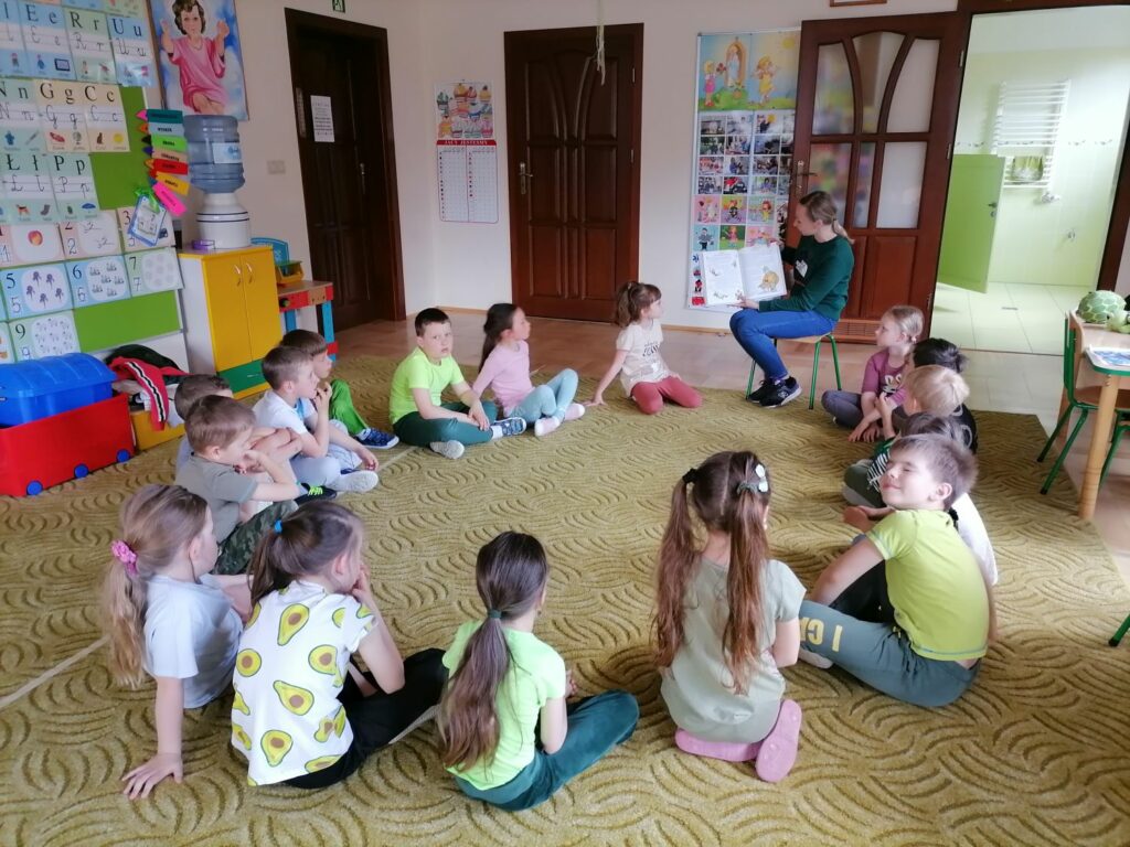Na zdjęciu widoczna jest grupa przedszkolaków siedzących na dywanie i słuchających bajki czytanej przez bibliotekarza.