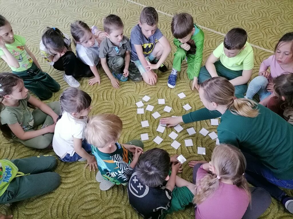 Na zdjęciu widoczna jest grupa przedszkolaków oraz bibliotekarz, którzy siedzą na dywanie i grają w memory.