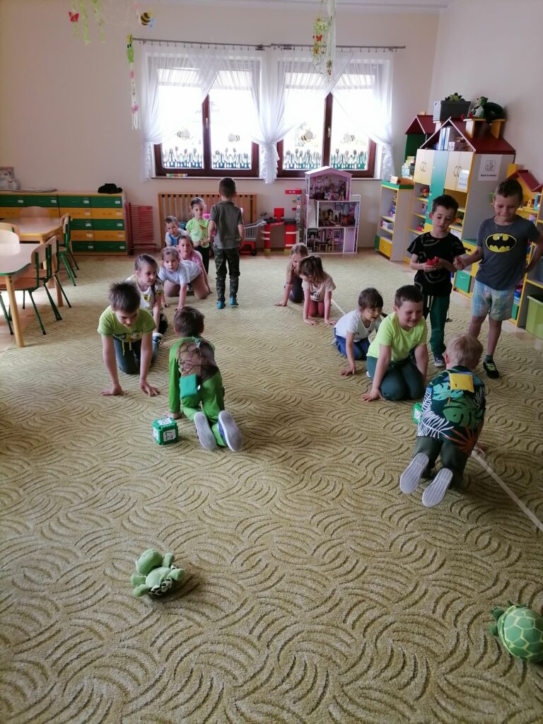 Na zdjęciu widoczna jest grupa przedszkolaków biorących udział w żółwiowych wyścigach polegających na przenoszeniu na plecach woreczków wokół przeszkód tak by ich nie upuścić.