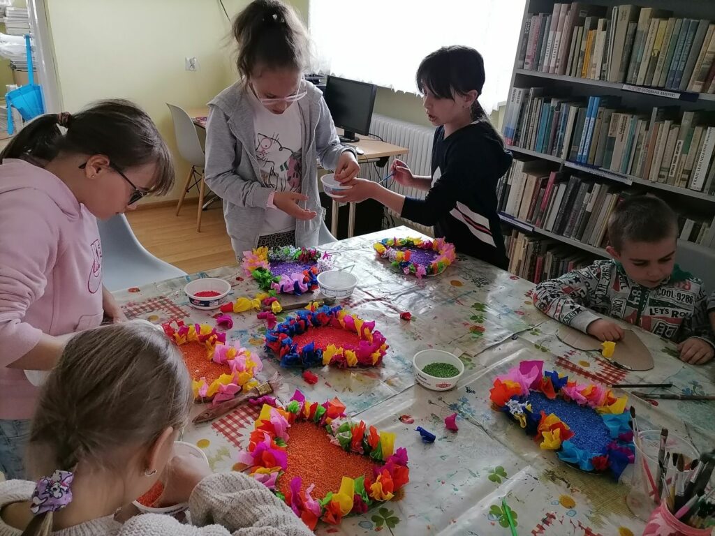 - Na zdjęciu widoczna jest grupka dzieci wykonujących prezent dla swoich mam. Dzieci siedzą przy stole i ozdabiają papierowe serca przy pomocy kolorowej bibuły i barwionego ryżu.