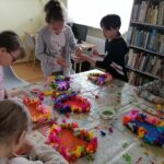 - Na zdjęciu widoczna jest grupka dzieci wykonujących prezent dla swoich mam. Dzieci siedzą przy stole i ozdabiają papierowe serca przy pomocy kolorowej bibuły i barwionego ryżu.