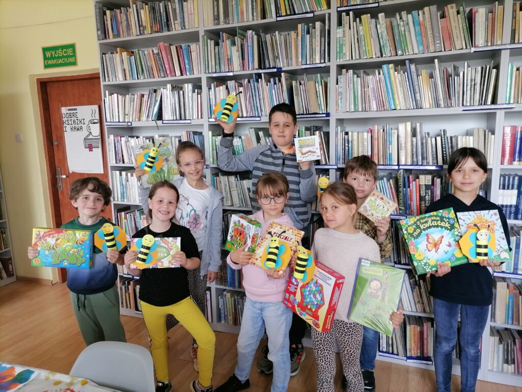 Na zdjęciu widoczna jest grupa dzieci uczestniczących w zajęciach z cyklu "Książka i gra - tematyczne rozgrywki gier planszowych" pozujących do wspólnego zdjęcia prezentujących wykorzystywane podczas zajęć książki, gry oraz wykonane przez siebie prace plastyczne.