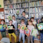 Na zdjęciu widoczna jest grupa dzieci uczestniczących w zajęciach z cyklu "Książka i gra - tematyczne rozgrywki gier planszowych" pozujących do wspólnego zdjęcia prezentujących wykorzystywane podczas zajęć książki, gry oraz wykonane przez siebie prace plastyczne.