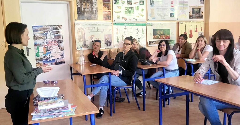 na zdjęciu widać stojącą bibliotekarkę oraz siedzące w ławkach osoby