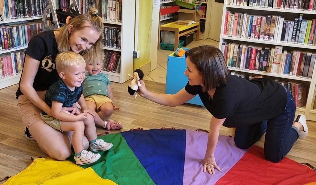 na zdjęciu widać dzieci siedzące na podłodze na kolorowej chuście oraz ich opiekunki