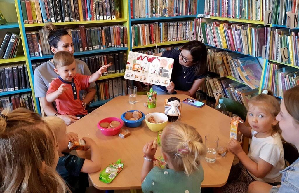 na zdjęciu widać dzieci i ich mamy siedzące wokół stolika i bibliotekarkę pokazującą im książkę