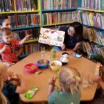 na zdjęciu widać dzieci i ich mamy siedzące wokół stolika i bibliotekarkę pokazującą im książkę