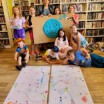 na zdjęciu widać dzieci w sali bibliotecznej stojące na tle regałów z książkami. dzieci trzymają prace plastyczne