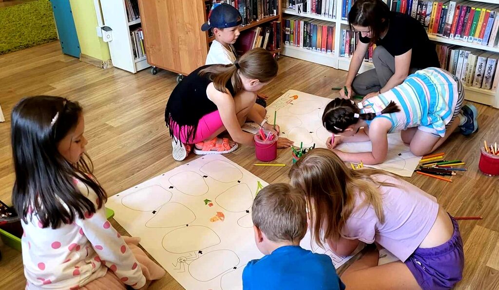 na zdjęciu widać rysujące na kartach rozłożonych na podłodze w sali bibliotecznej