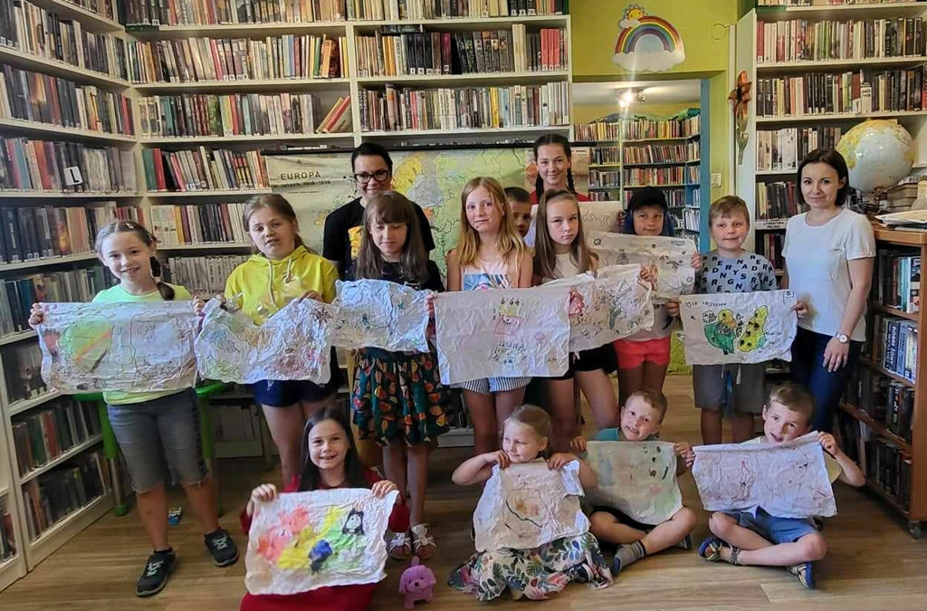 na zdjęciu widać salę biblioteczną, regały z książkami i dzieci wraz z bibliotekarkami