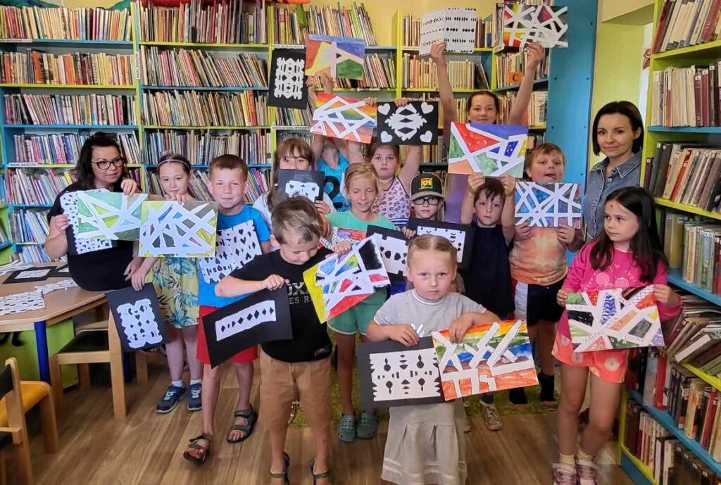 na zdjęciu widać dzieci w sali bibliotecznej trzymające obrazki przez siebie wykonane