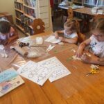 na zdjęciu widać salę biblioteczną, dzieci siedzą przy stole wykonują pracę plastyczną