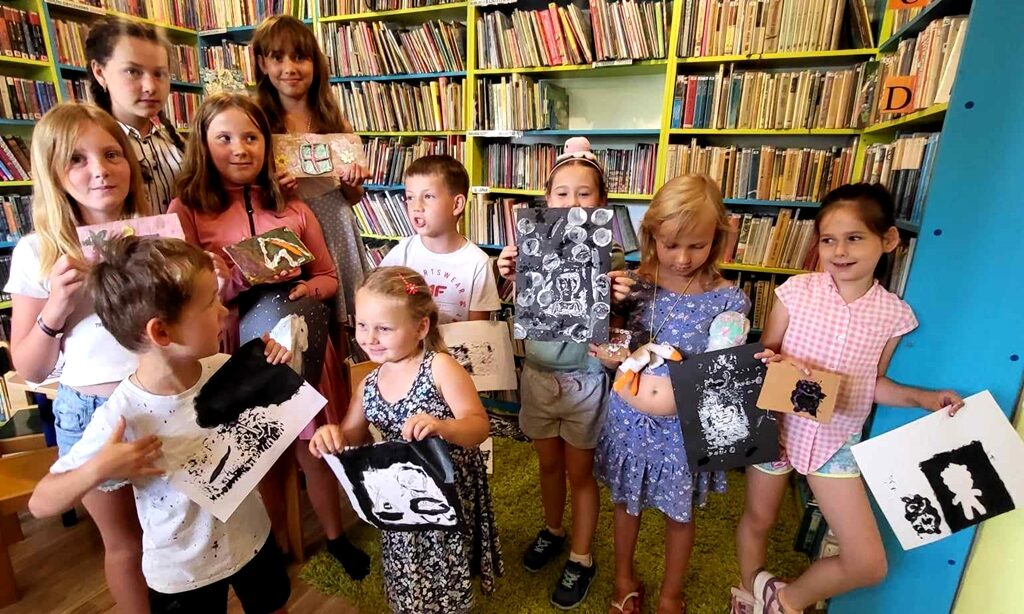 Na zdjęciu widać dzieci stojące na tle regału z książkami. Dzieci trzymają rysunki.