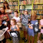 Na zdjęciu widać dzieci stojące na tle regału z książkami. Dzieci trzymają rysunki.