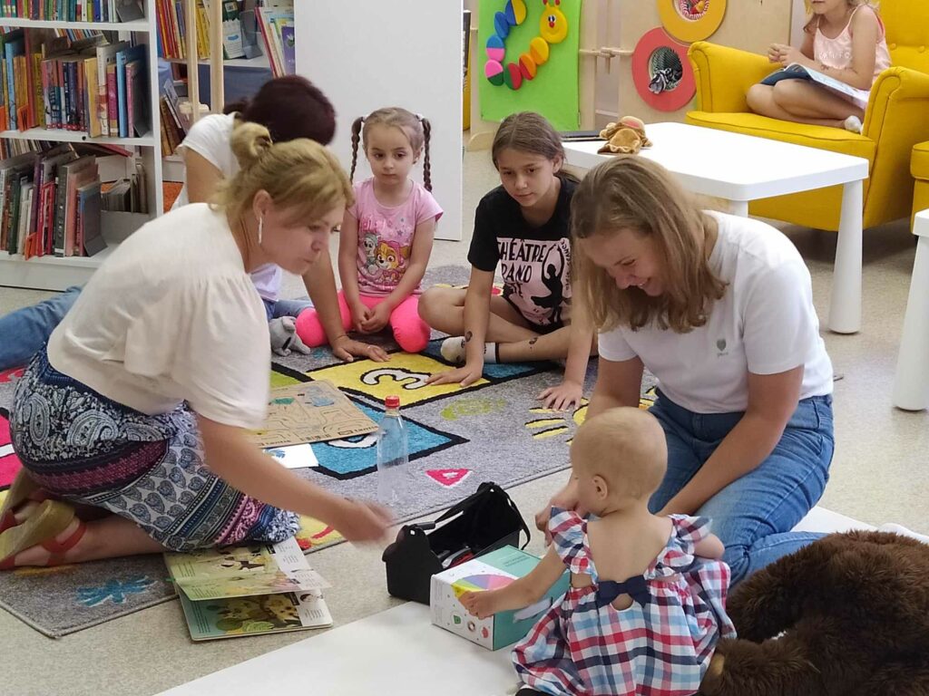 Na dywanie siedzą dzieci wraz z mamami i panią bibliotekarką i słuchająjak panibibliotekarka czyta książeczkę.
