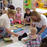 Na dywanie siedzą dzieci wraz z mamami i panią bibliotekarką i słuchająjak panibibliotekarka czyta książeczkę.
