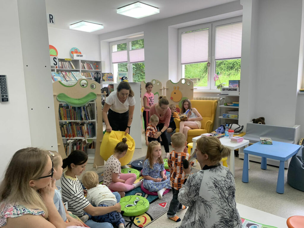 Na zdjęciu sala biblioteczna dla dzieci a w niej na podłodze siedzą dzieci w wieku trzy cztery lata wraz ze swoimi mamami. Wszyscy uczestnicza w zajęciach przygotowanych przez bibliotekarkę, która wraz z nimi siedzi na podłodze.
