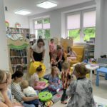 Na zdjęciu sala biblioteczna dla dzieci a w niej na podłodze siedzą dzieci w wieku trzy cztery lata wraz ze swoimi mamami. Wszyscy uczestnicza w zajęciach przygotowanych przez bibliotekarkę, która wraz z nimi siedzi na podłodze.