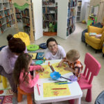 Na zdjęciu sala biblioteczna dla dzieci. Dzieci w wieku 2-4 lata siedzą przy stoliczkach i wykonują prace plastyczne wraz z rodzicami