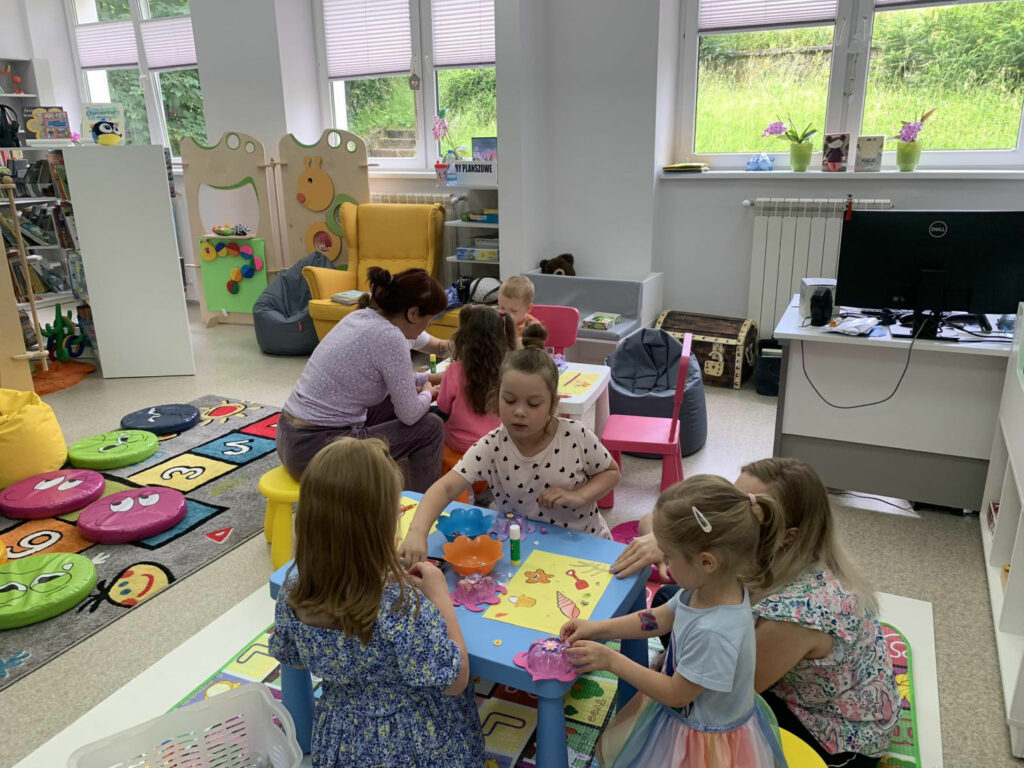 Na zdjęciu sala biblioteczna dla dzieci. Dzieci w wieku 2-4 lata siedzą przy stoliczkach i wykonują prace plastyczne wraz z rodzicami