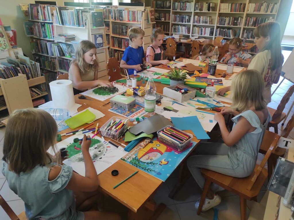na zdjęciu widać salę biblioteczną, przy stole siedzą dzieci, wykonują prace plastyczne