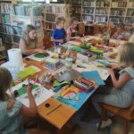 na zdjęciu widać salę biblioteczną, przy stole siedzą dzieci, wykonują prace plastyczne